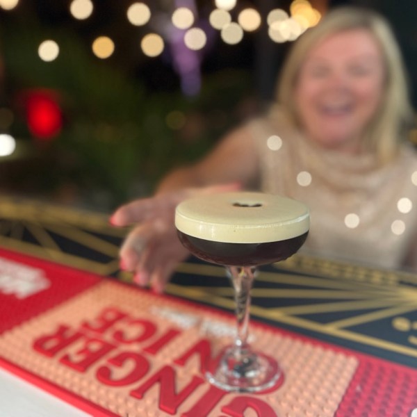 Woman reaching for a cocktail with a creamy top at a bar, blurred festive lights in background.