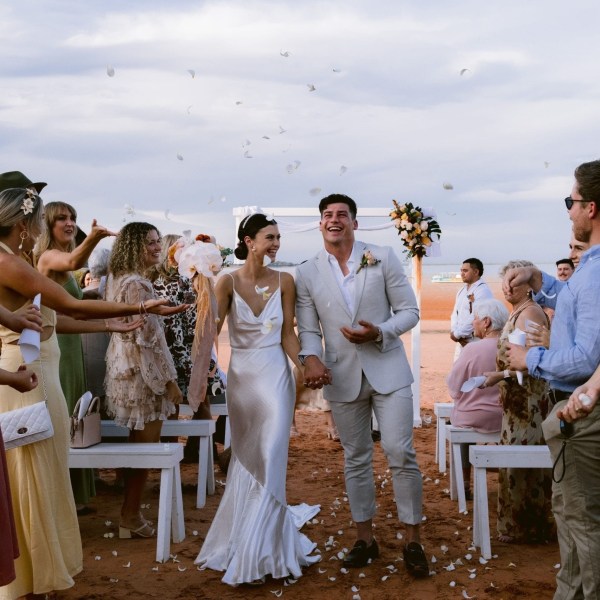 Bride and groom walk down aisle, guests toss petals, outdoor wedding on a sandy beach.