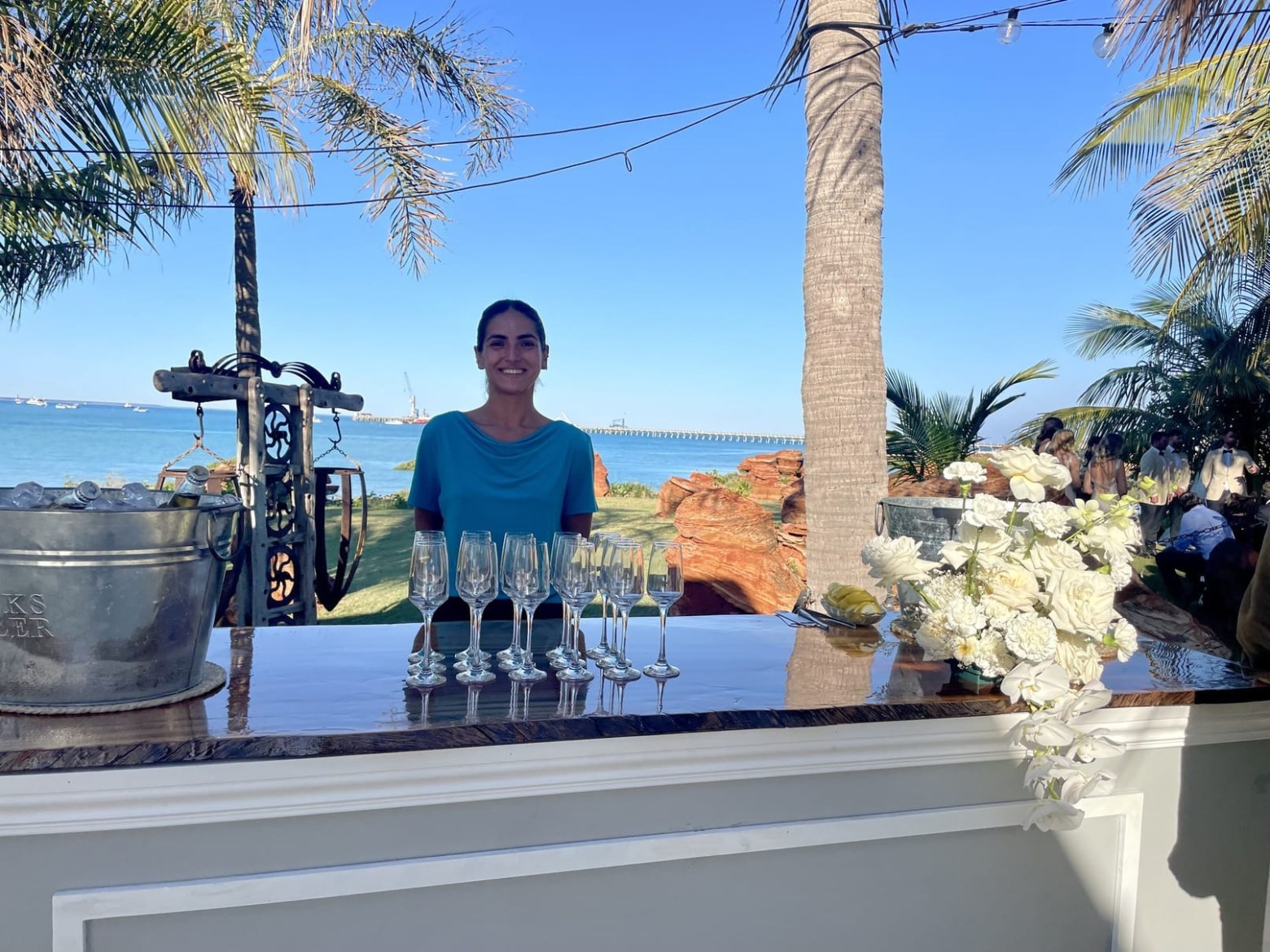 Smiling woman behind outdoor bar with champagne flutes, ocean view, and palm trees.