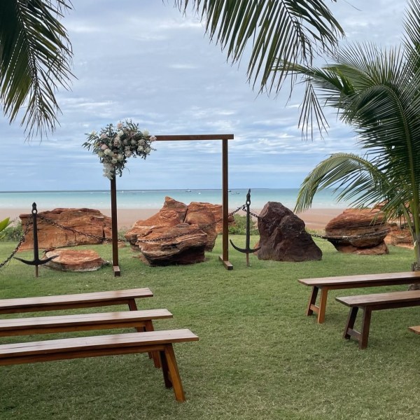 Outdoor wedding setup with benches, an arch, and a beach view under palm trees.