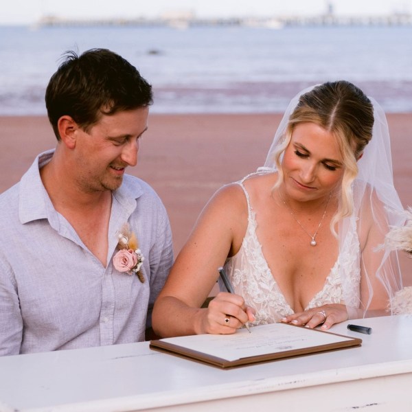Bride signs document with groom watching on beach, floral arrangement nearby.