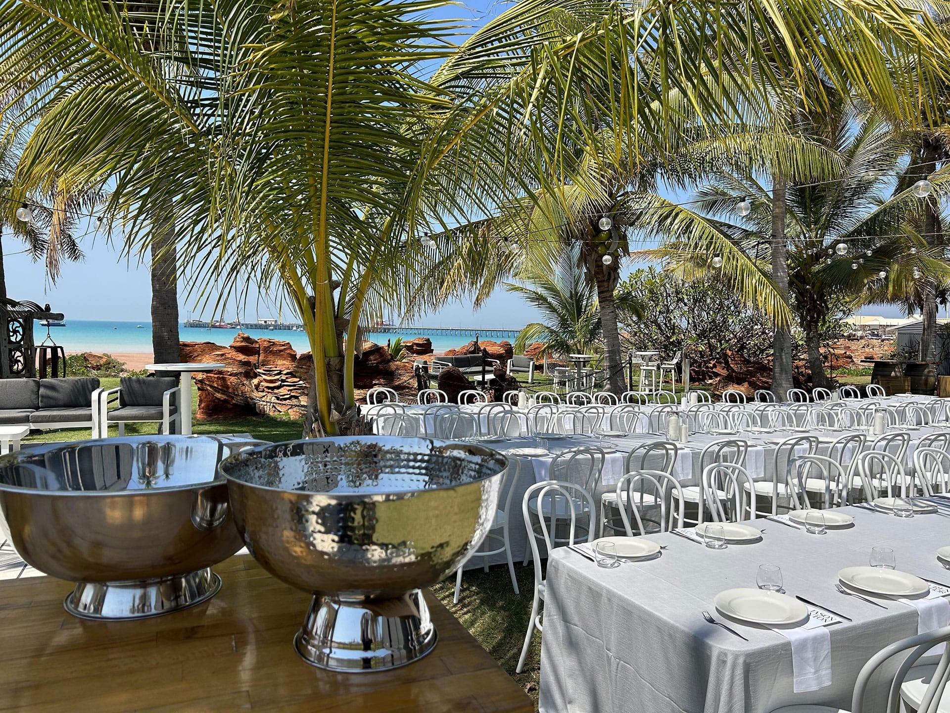 Outdoor dining setup with white tables and chairs under palm trees near a beach on a sunny day.