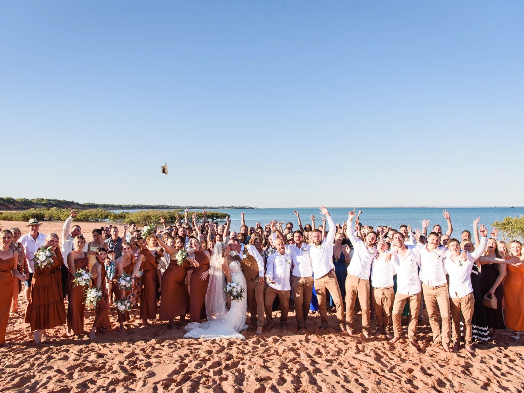 Large wedding party cheering on a sunny beach with blue sky and ocean backdrop.