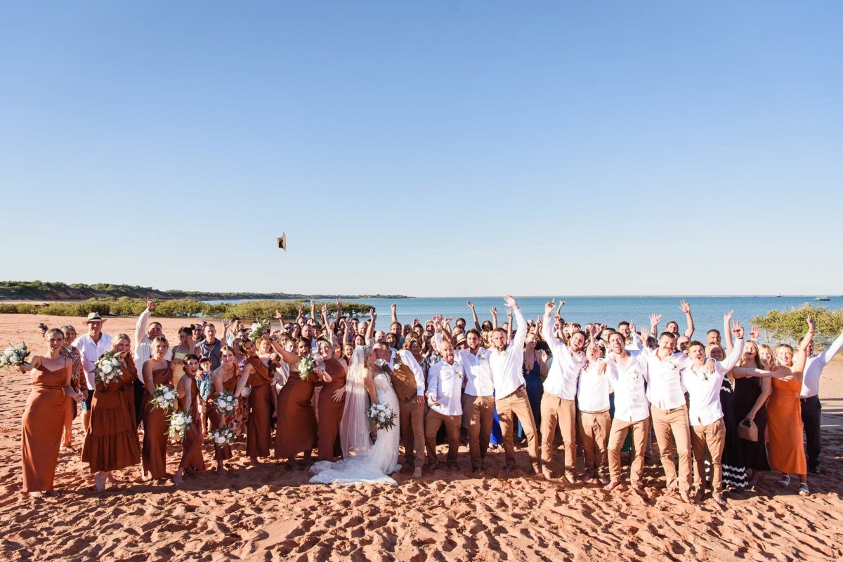 Large wedding party cheering on a sunny beach with blue sky and ocean backdrop.