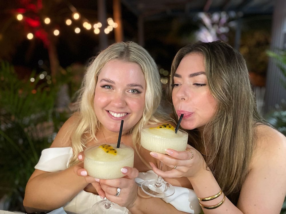Two women smiling and drinking cocktails with black straws in a nighttime setting.