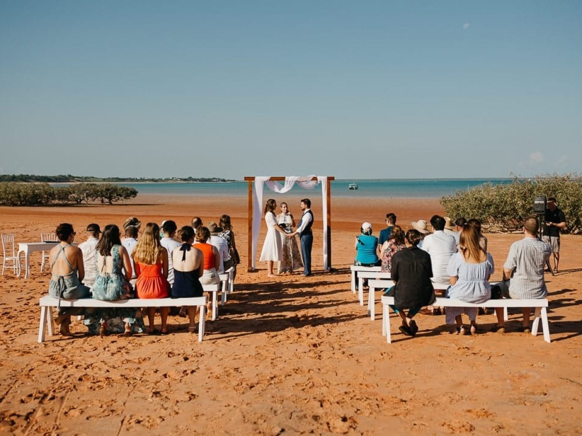 Beach wedding ceremony with guests seated on benches facing the water.