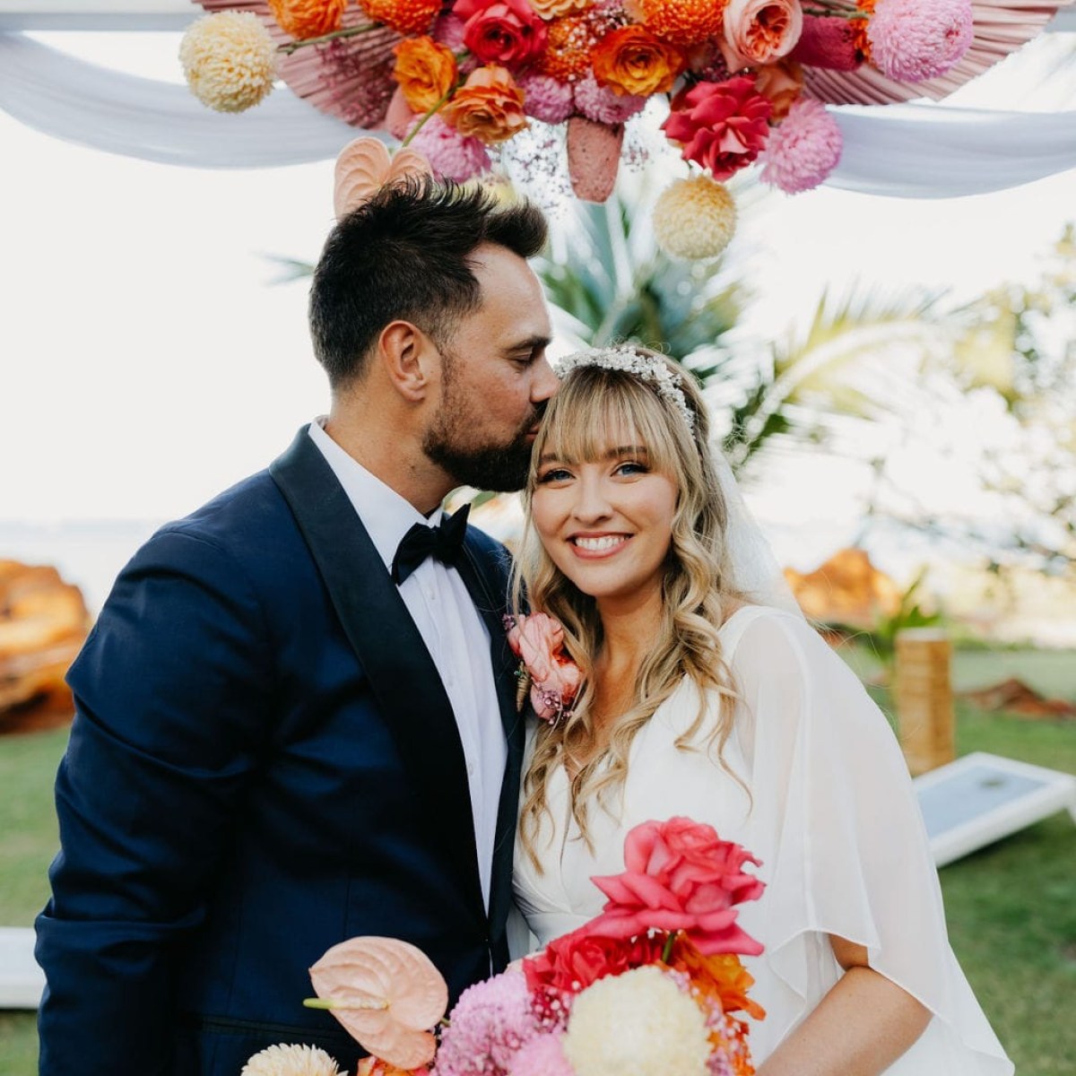 Bride and groom smiling, groom kisses bride's head, holding colorful bouquet in outdoor setting.