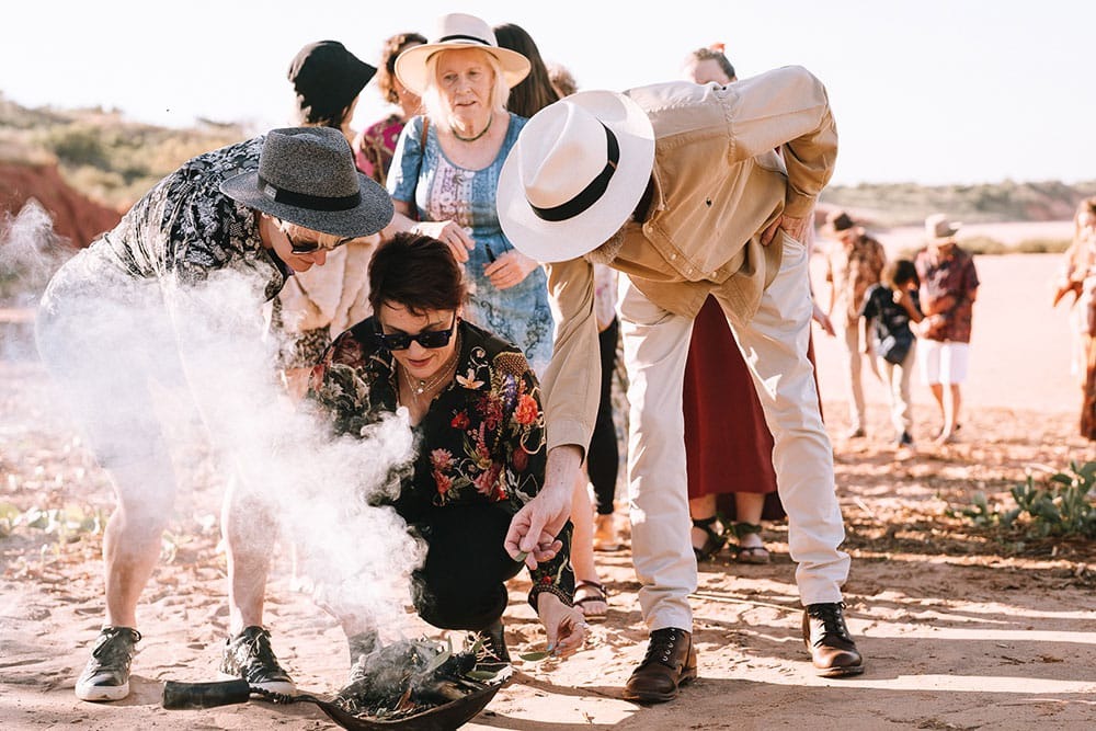 Group of people outdoors observing a smoking bowl on the ground, wearing hats and casual clothing.