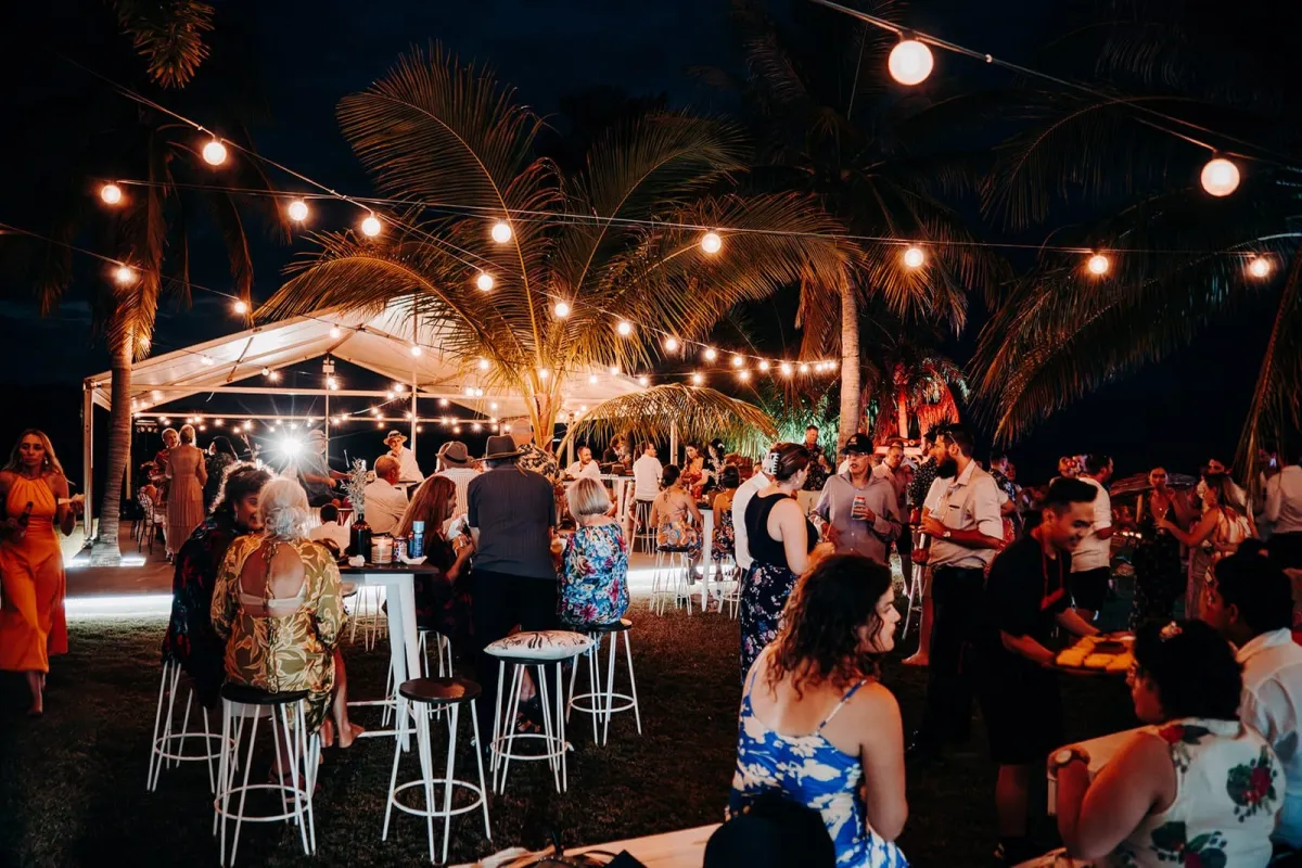Outdoor evening gathering with people under string lights and palm trees.