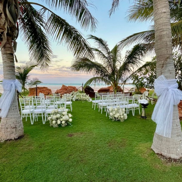 Outdoor wedding setup with white chairs, flowers, and palm trees by the ocean.