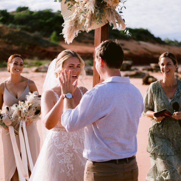 Wedding ceremony on beach with bride smiling, holding groom's hands, bridesmaids nearby, and officiant.
