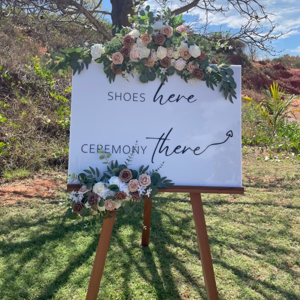 Wedding sign with flowers reads 'Shoes here, Ceremony there' on an easel outdoors.