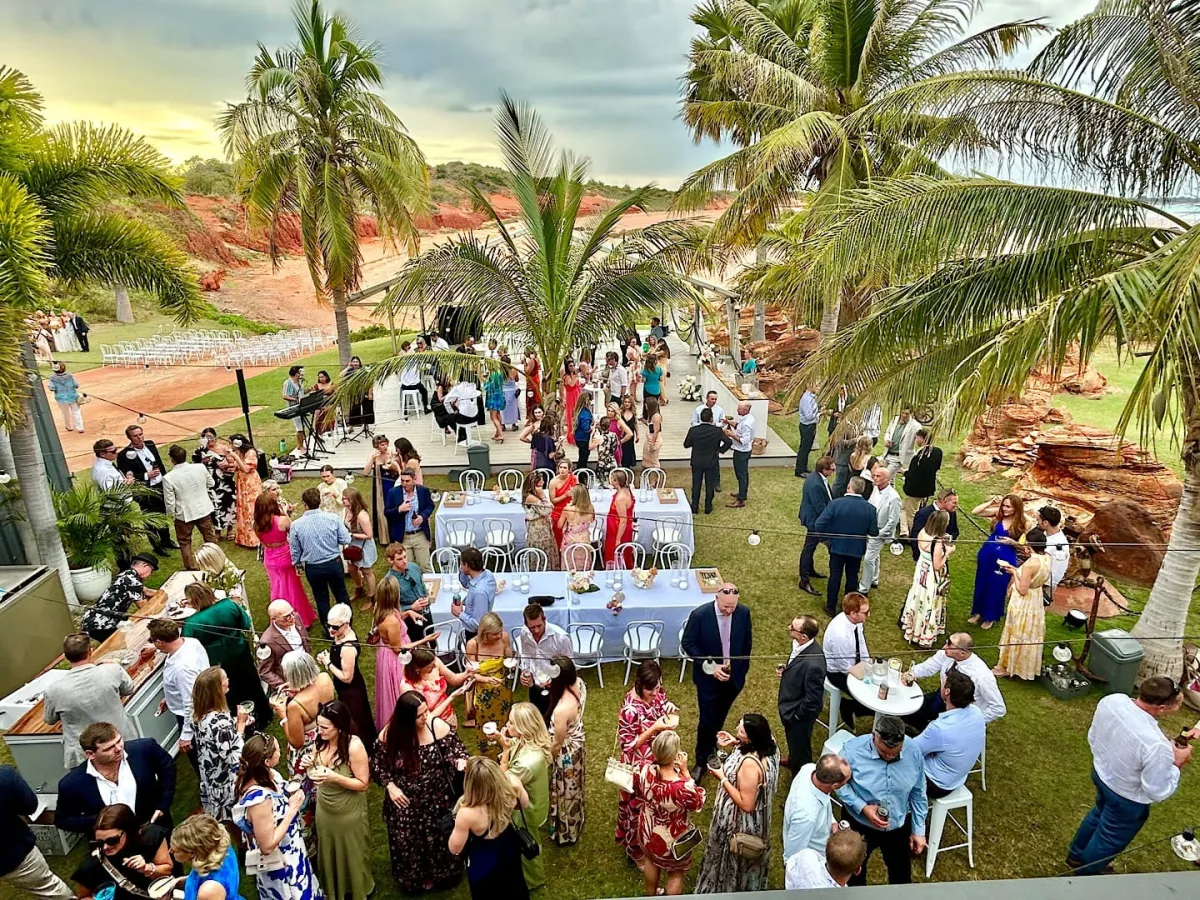 Outdoor gathering with people mingling under palm trees at a scenic venue.