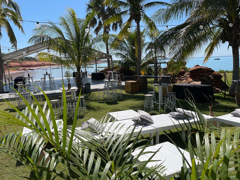 Outdoor event area with white tables, chairs, palm trees, and ocean view under clear blue sky.