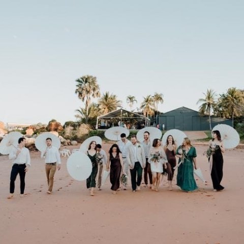 People on a beach holding white parasols, with greenery in the background.