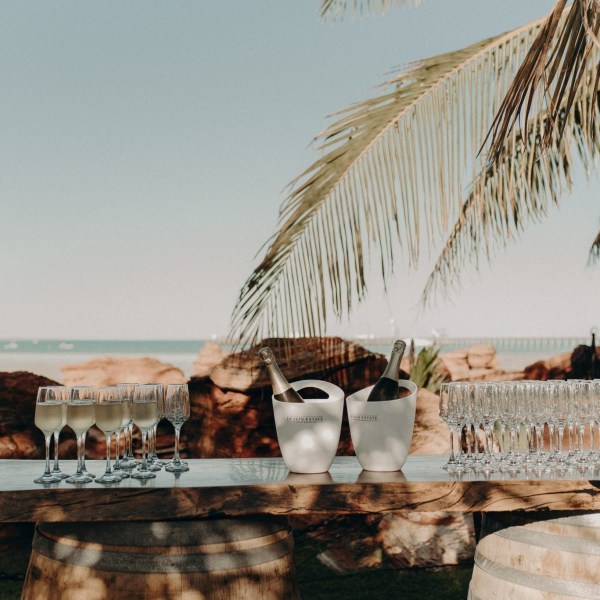 Outdoor setup with champagne flutes, bottles in ice buckets on a wooden table, and a palm tree.