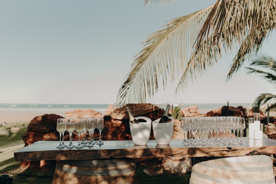 Outdoor setup with champagne flutes, bottles in ice buckets on a wooden table, and a palm tree.