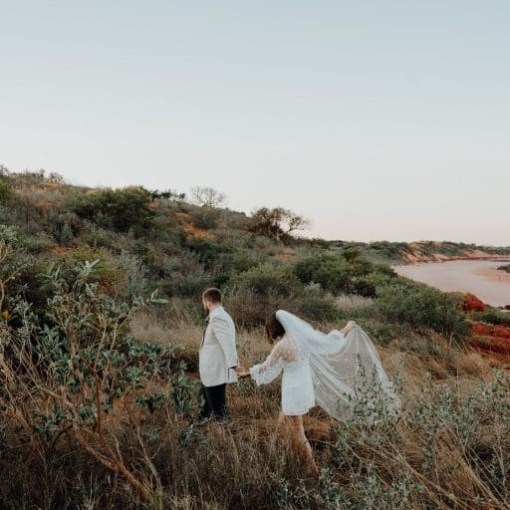 Bride and groom walking through grassy field by a beach at sunset.