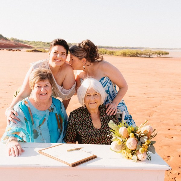 Four women smiling at a table on a sandy beach, with one holding a bouquet.