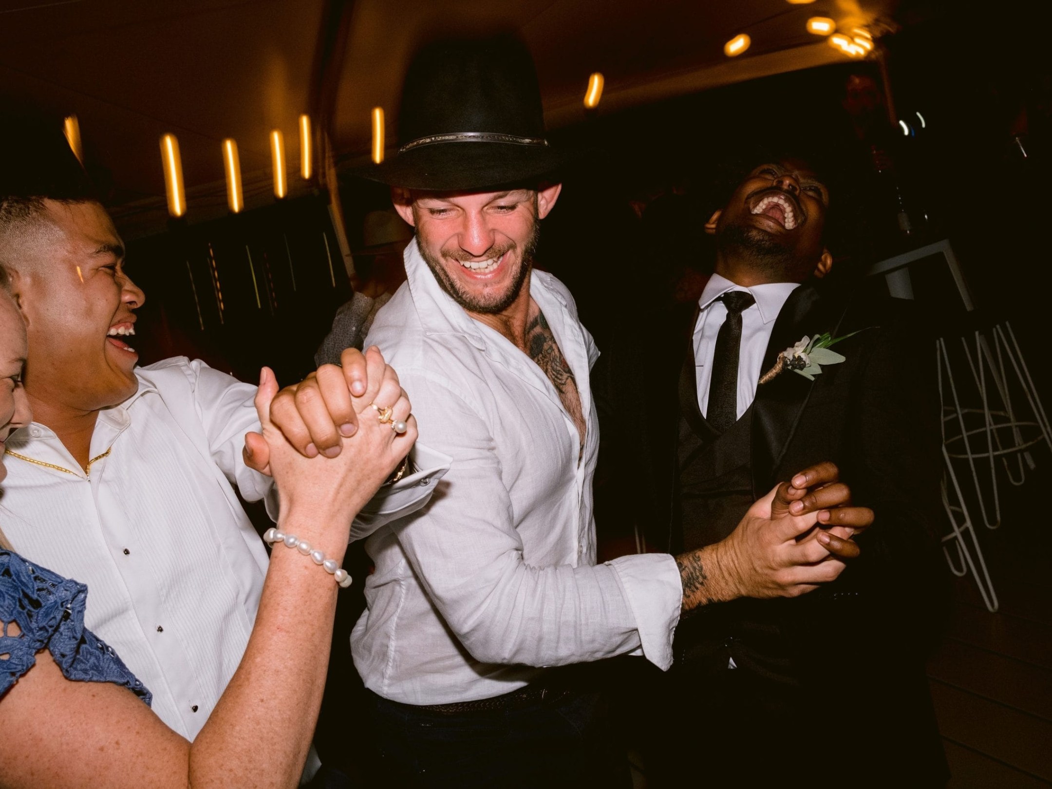 People laughing and dancing at a party, one wearing a hat and another in a suit with a boutonniere.