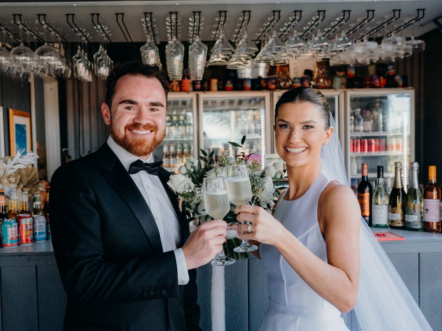 Bride and groom smiling, holding champagne glasses at a bar with bottles in the background.
