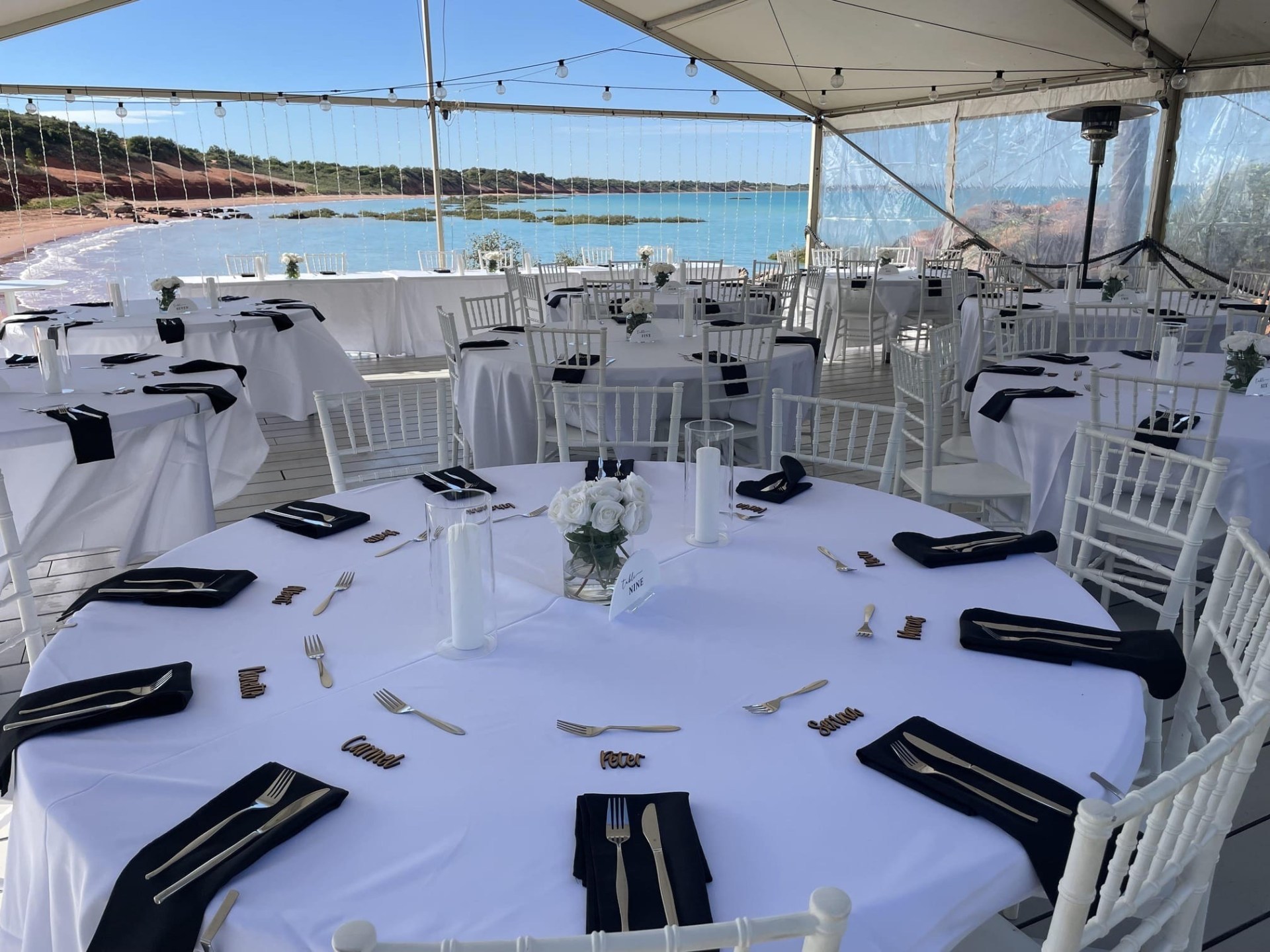 Outdoor wedding reception setup with white tables, chairs, black napkins, and ocean view.