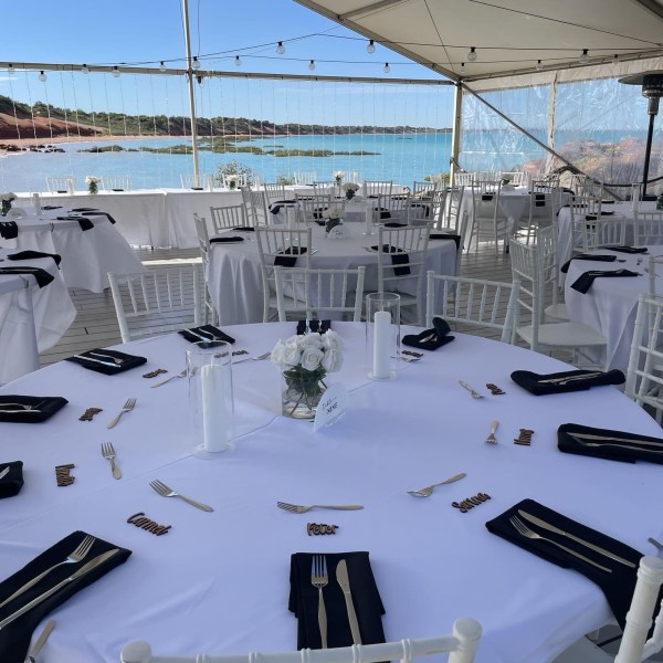 Outdoor wedding reception setup with white tables, chairs, black napkins, and ocean view.