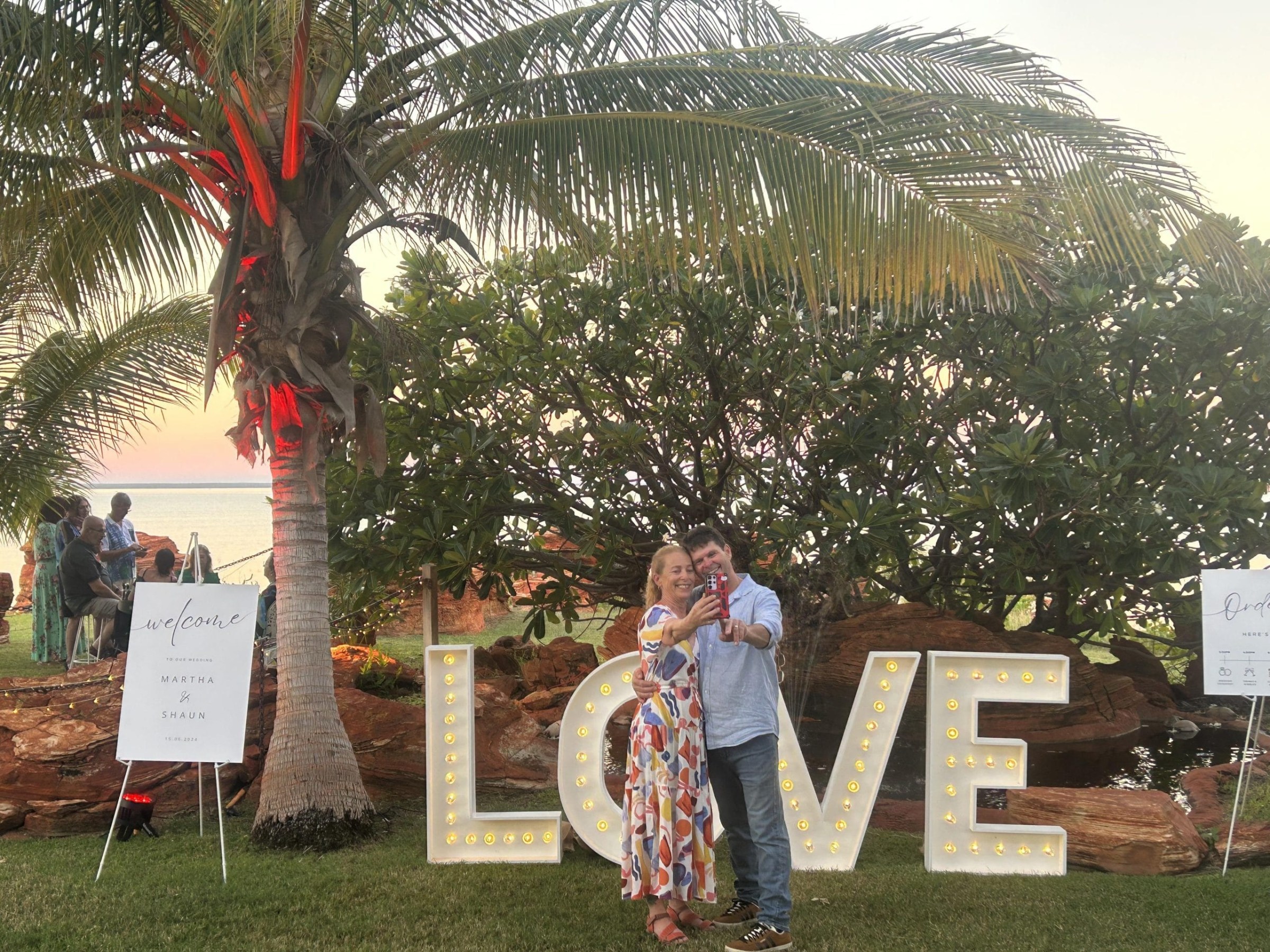 A couple taking a selfie in front of large illuminated letters spelling LOVE under a palm tree.