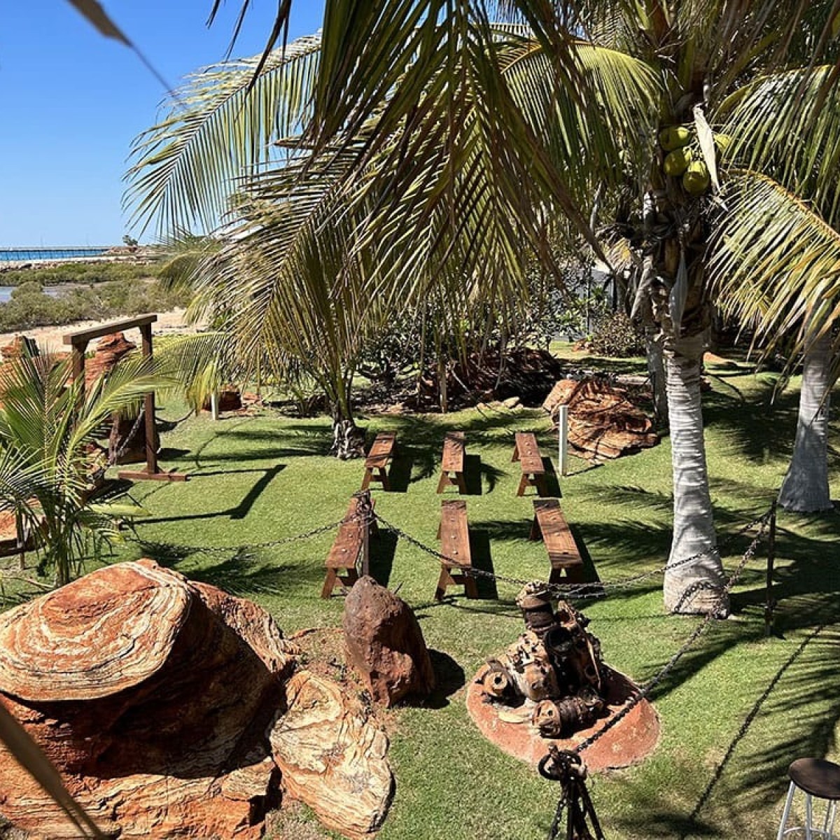 Tropical garden with palm trees, wooden benches, and rocky formations near a beach with ocean and blue sky.