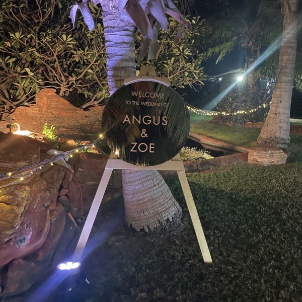 Night wedding welcome sign on an easel under a palm tree, with lights and foliage.