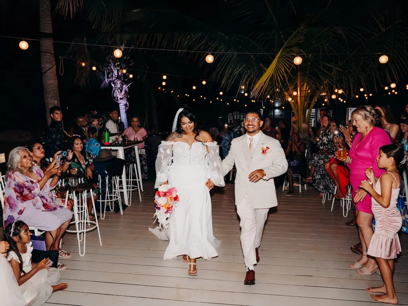Bride and groom walking hand in hand, surrounded by cheering guests under string lights at night.