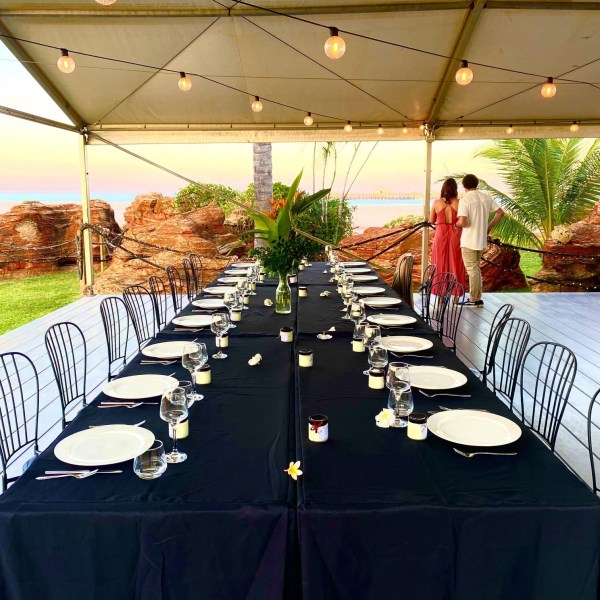 Outdoor dining setup with long table, black tablecloth, empty plates, and string lights under a tent.