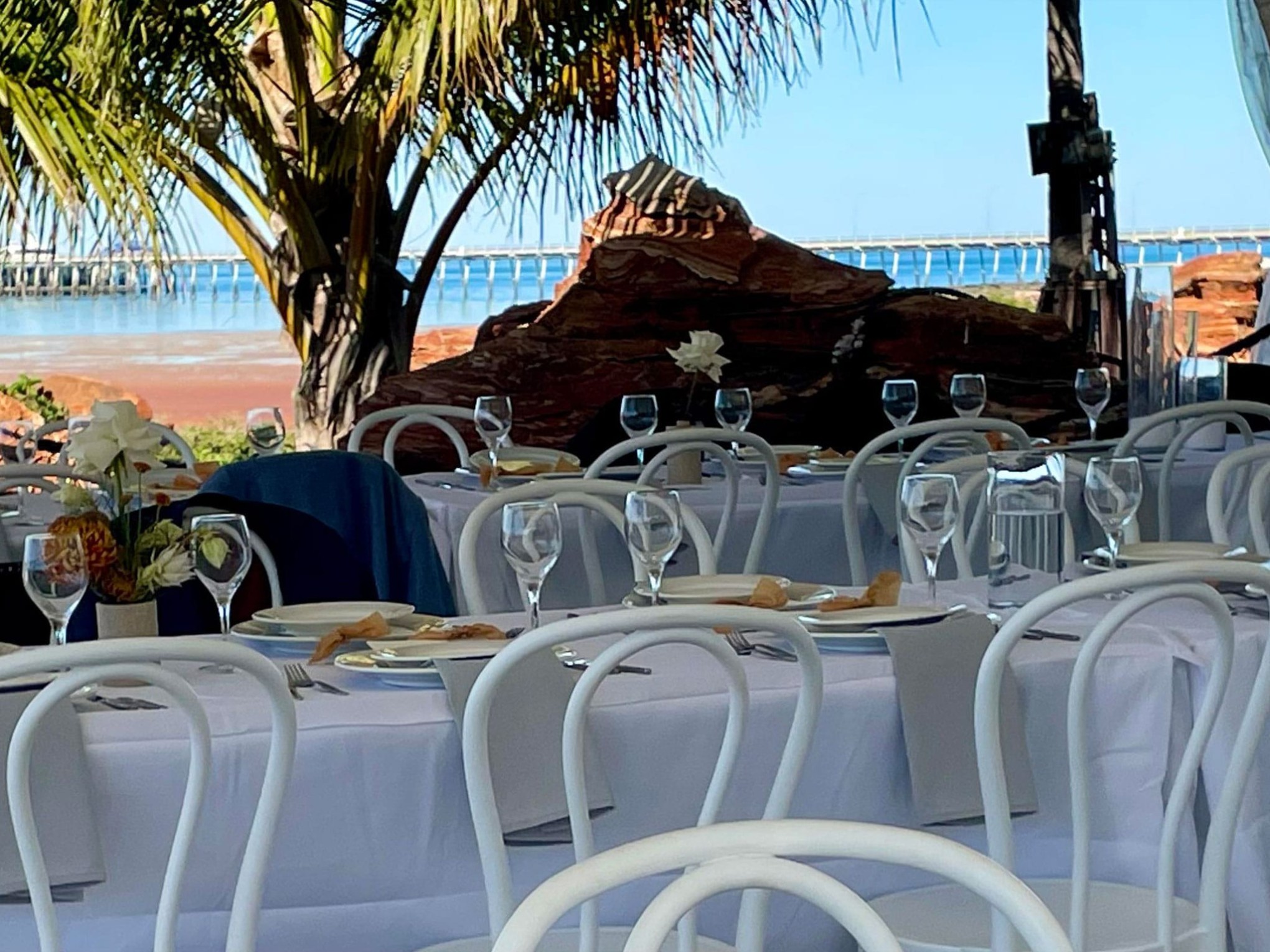 Outdoor dining setup with white tablecloths and plates near beach, palm trees, and a pier.