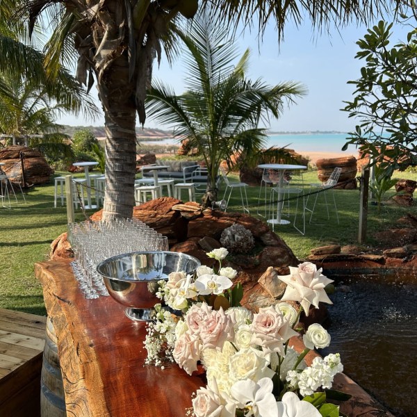 Outdoor setting with flowers, palm trees, and ocean view; glasses on a wooden table.
