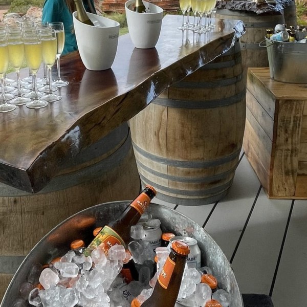 Outdoor bar with drinks on wooden barrel table, scenic view of red rocks and ocean, person standing behind.
