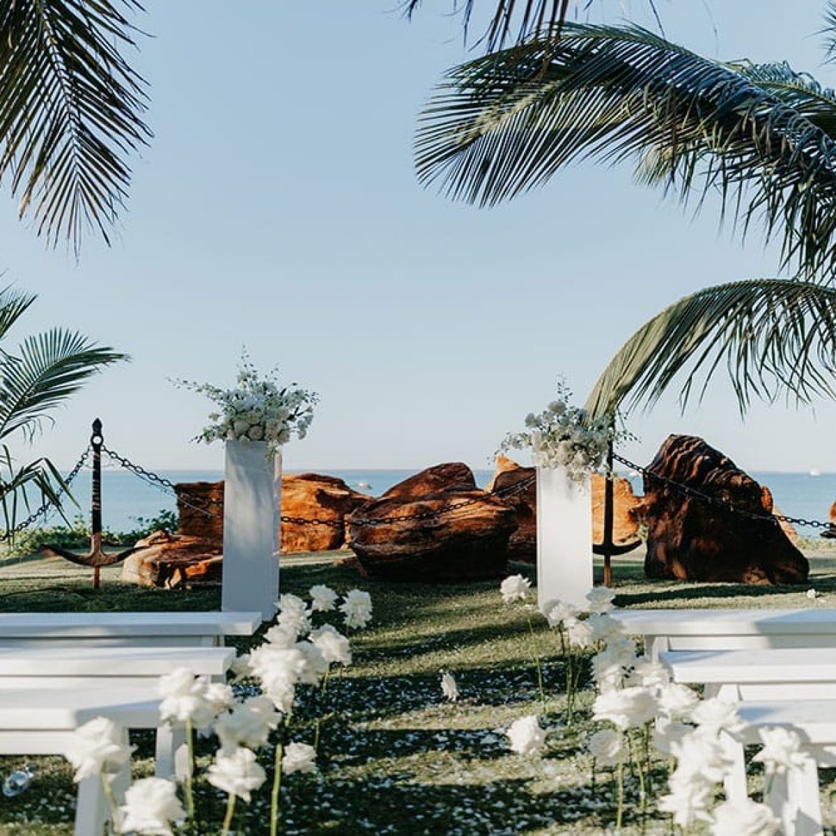 Outdoor wedding setup by the beach with palm trees and white benches.