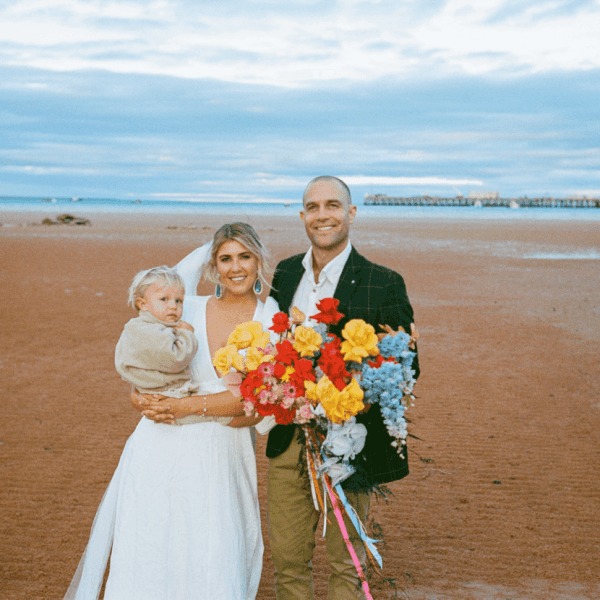 Three people on a beach, two adults and a child, smiling and holding colorful flowers.