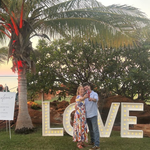Couple takes selfie by large 'LOVE' sign, palm trees, ocean backdrop.