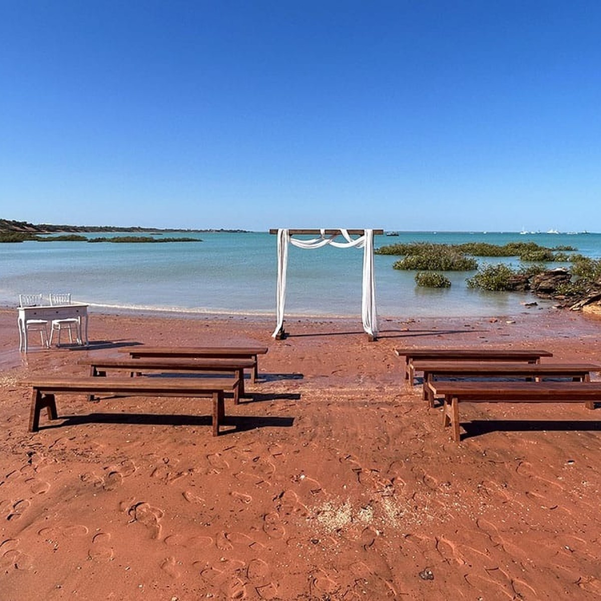 Beach wedding setup with benches, white arch, and chairs on red sand near calm water.