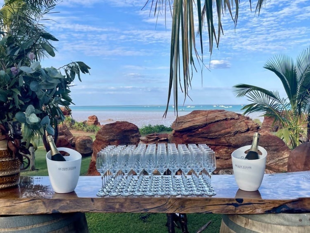 Outdoor bar setup with champagne bottles, glasses, and flowers by a beach with rocks and palm trees.