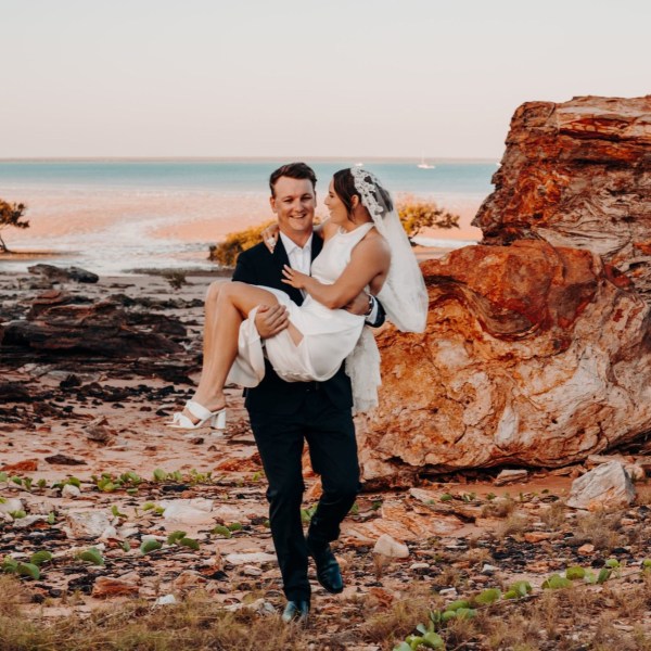Groom carrying bride on beach with rocky formations and ocean background at sunset.