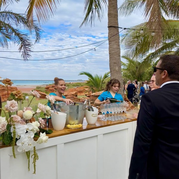 Outdoor bar with bartenders under palm trees, ocean in background, and guests mingling.