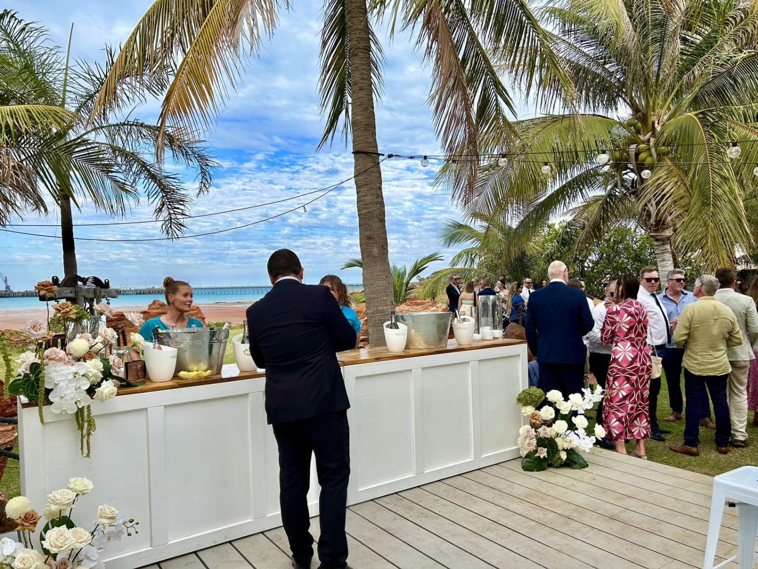 Outdoor event with people at a white bar under palm trees on a sunny day.