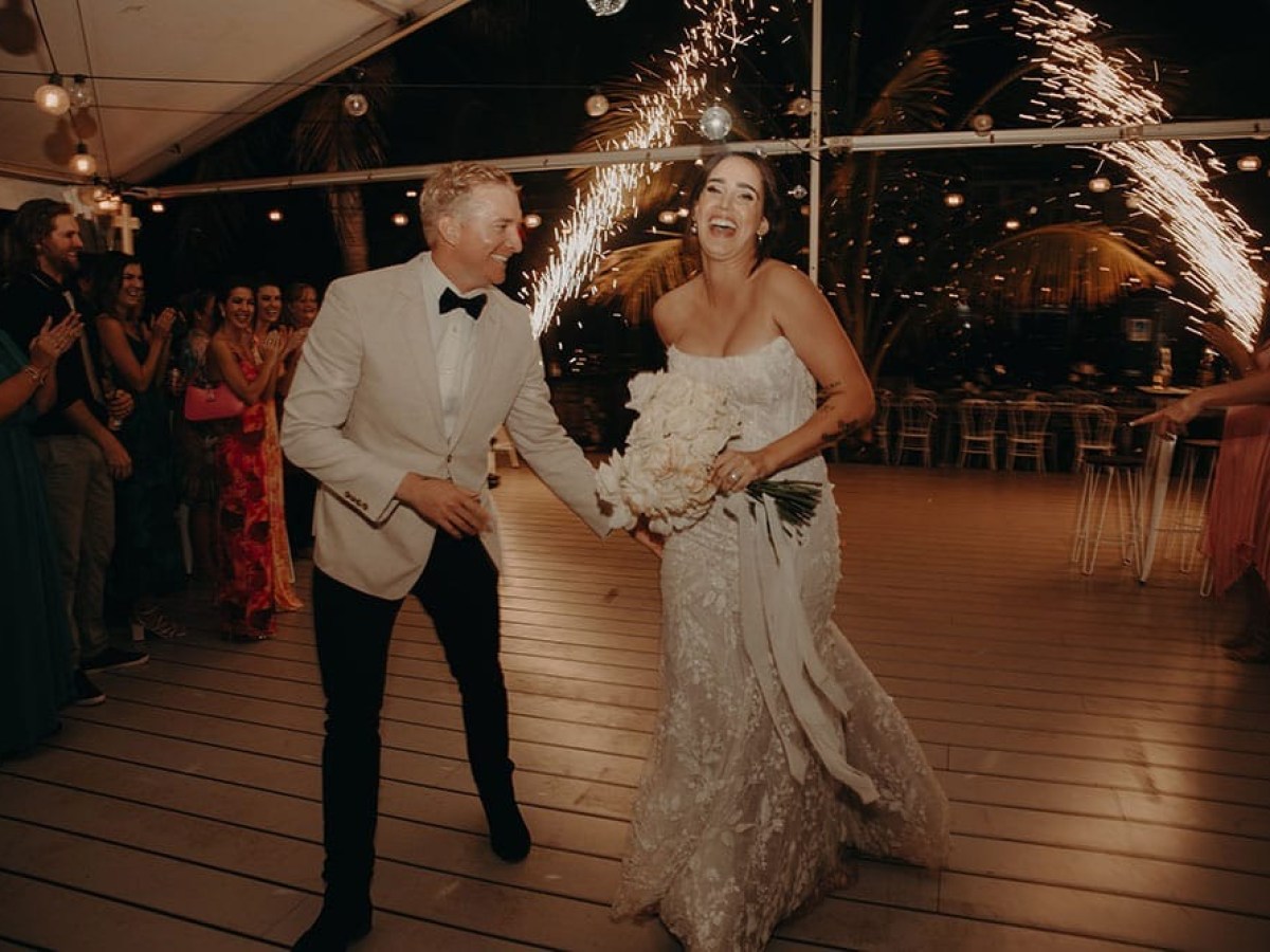Bride and groom smiling and holding hands on dance floor with sparklers in the background.