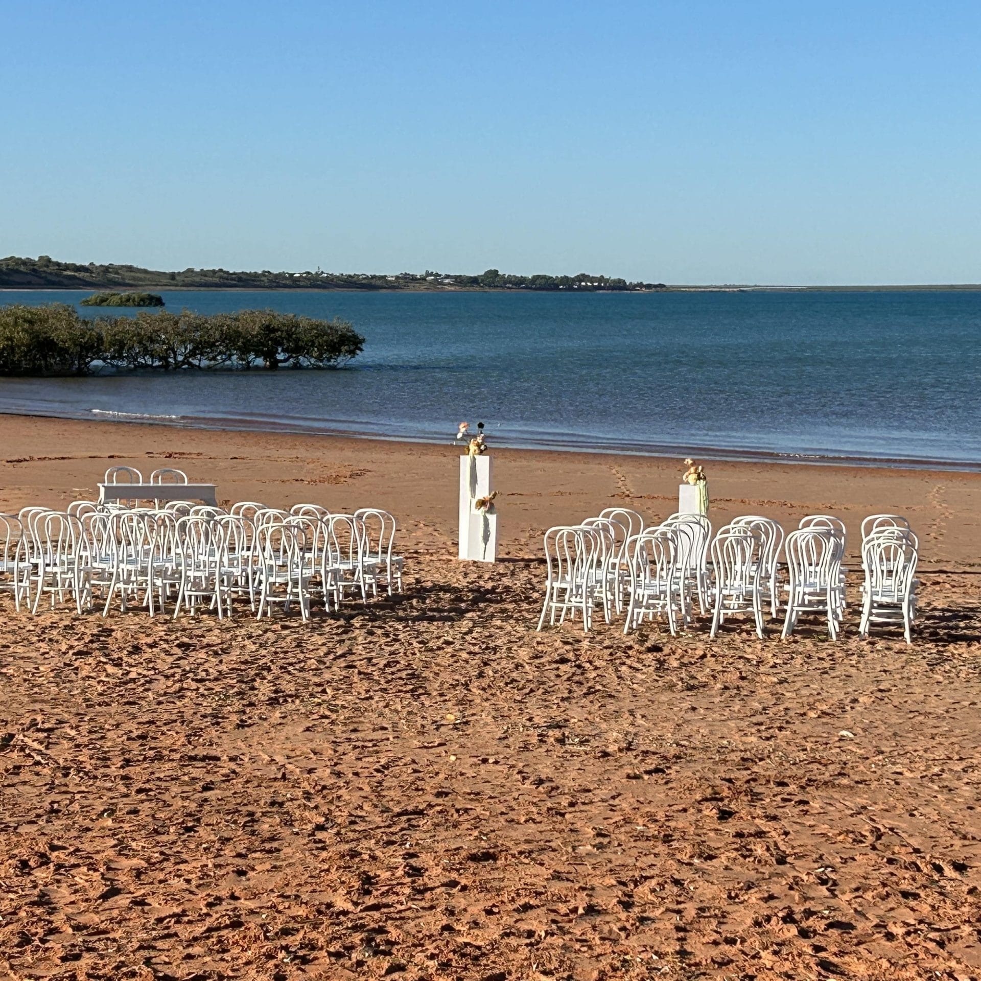 Rows of white chairs set up on a sandy beach, facing the ocean, with two small podiums.