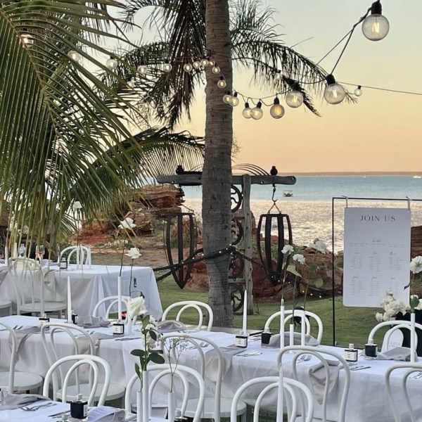 Outdoor dining setup with white tables and chairs, palm trees, and a sea view at sunset.