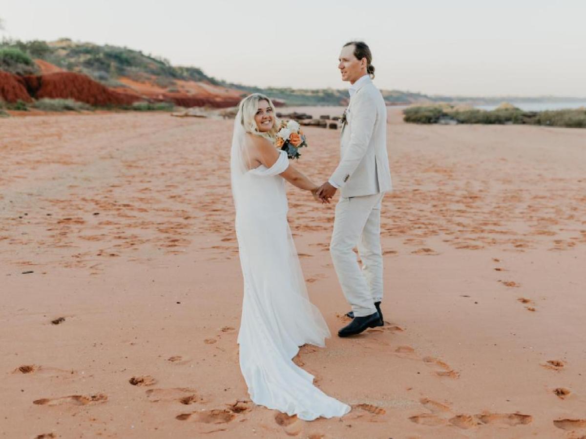 Bride and groom in white attire holding hands on a sandy beach.