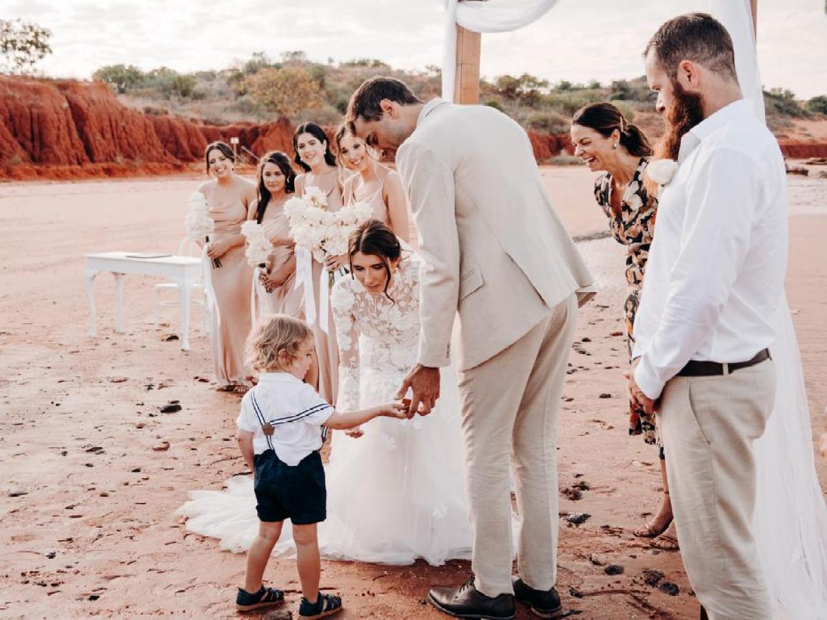 Bride and groom at a beach wedding greet a young boy with a group of bridesmaids smiling nearby.