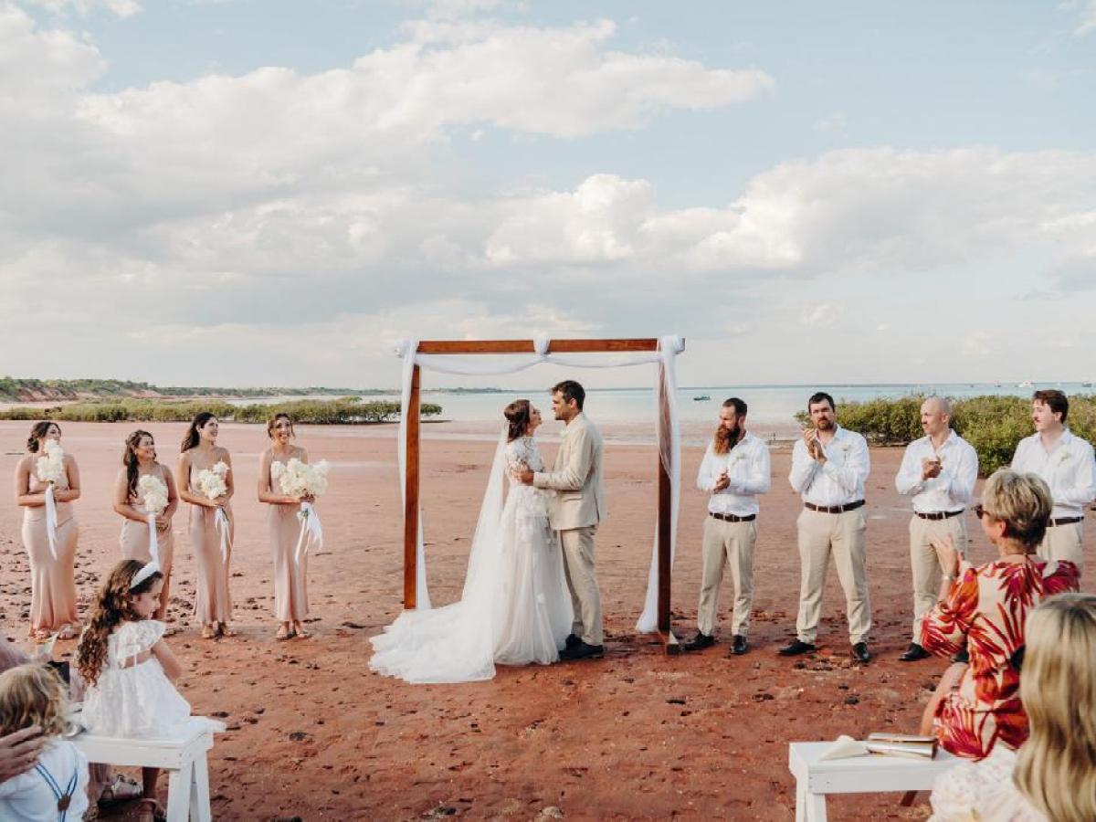Beach wedding ceremony with a couple under an arch, surrounded by bridesmaids and groomsmen.