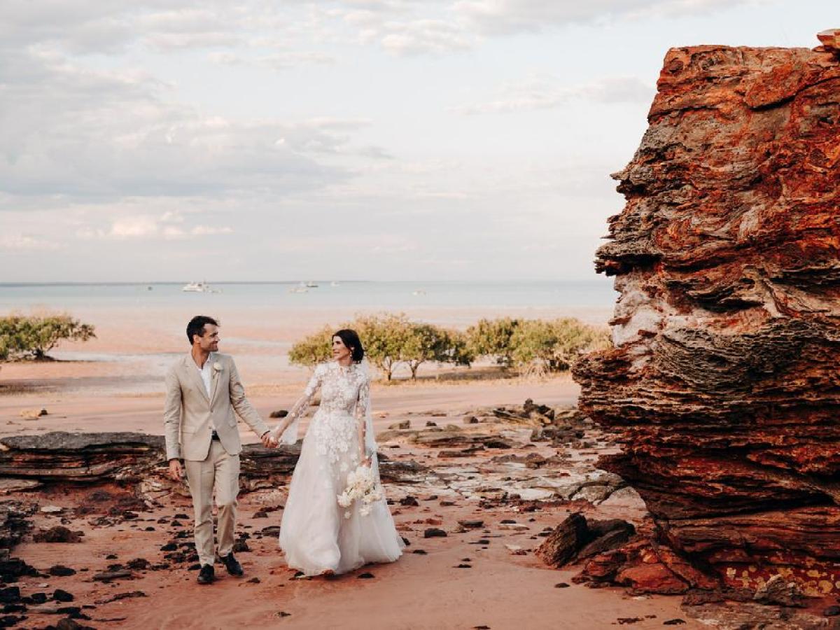 Couple in wedding attire holding hands on a rocky beach with large boulder and ocean view.